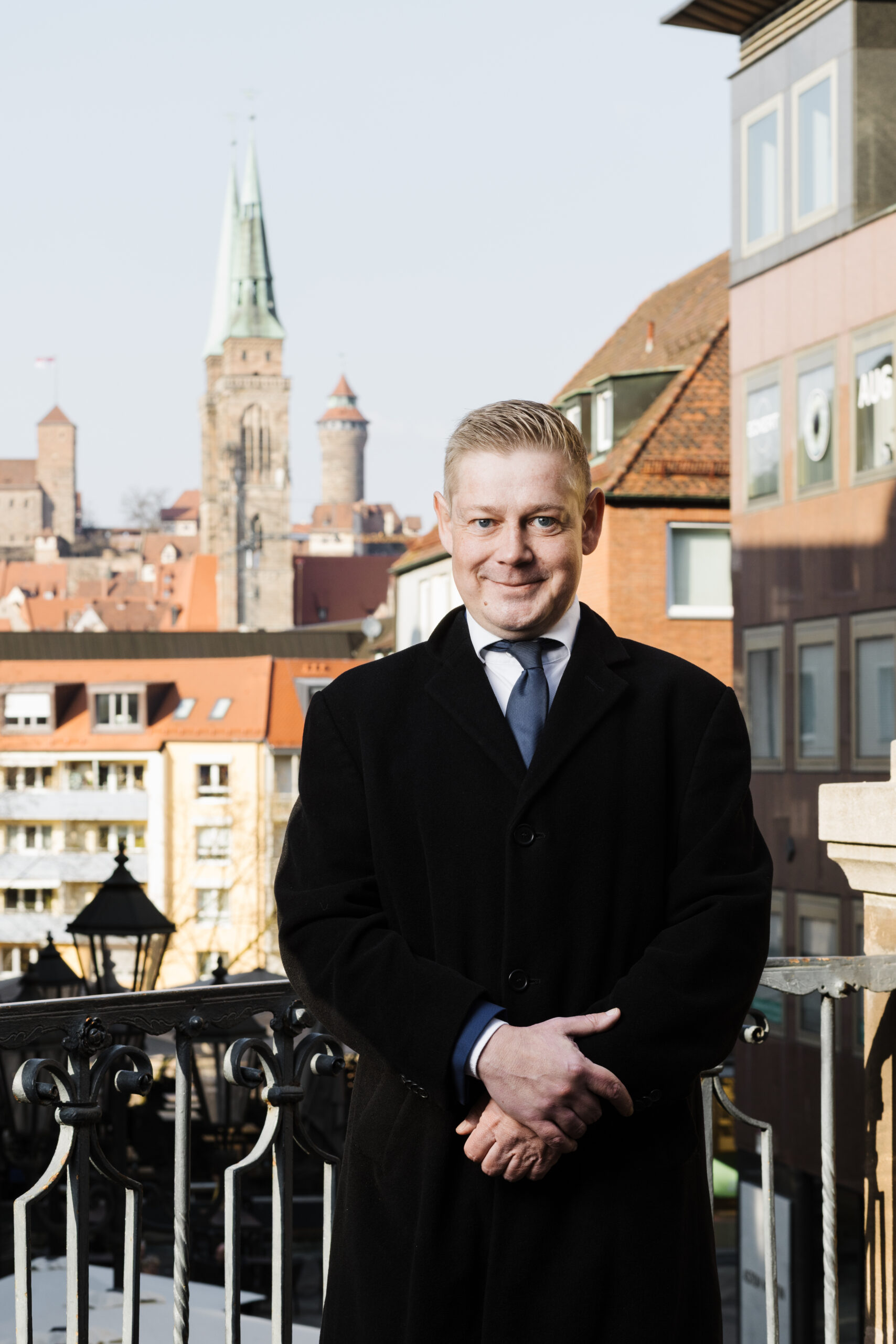 Fotografie Rechtsanwalt Kevin Nowack in schwarzem Mantel, Hemd und Krawatte, im Hintegrund Gebäude von Nürnberg