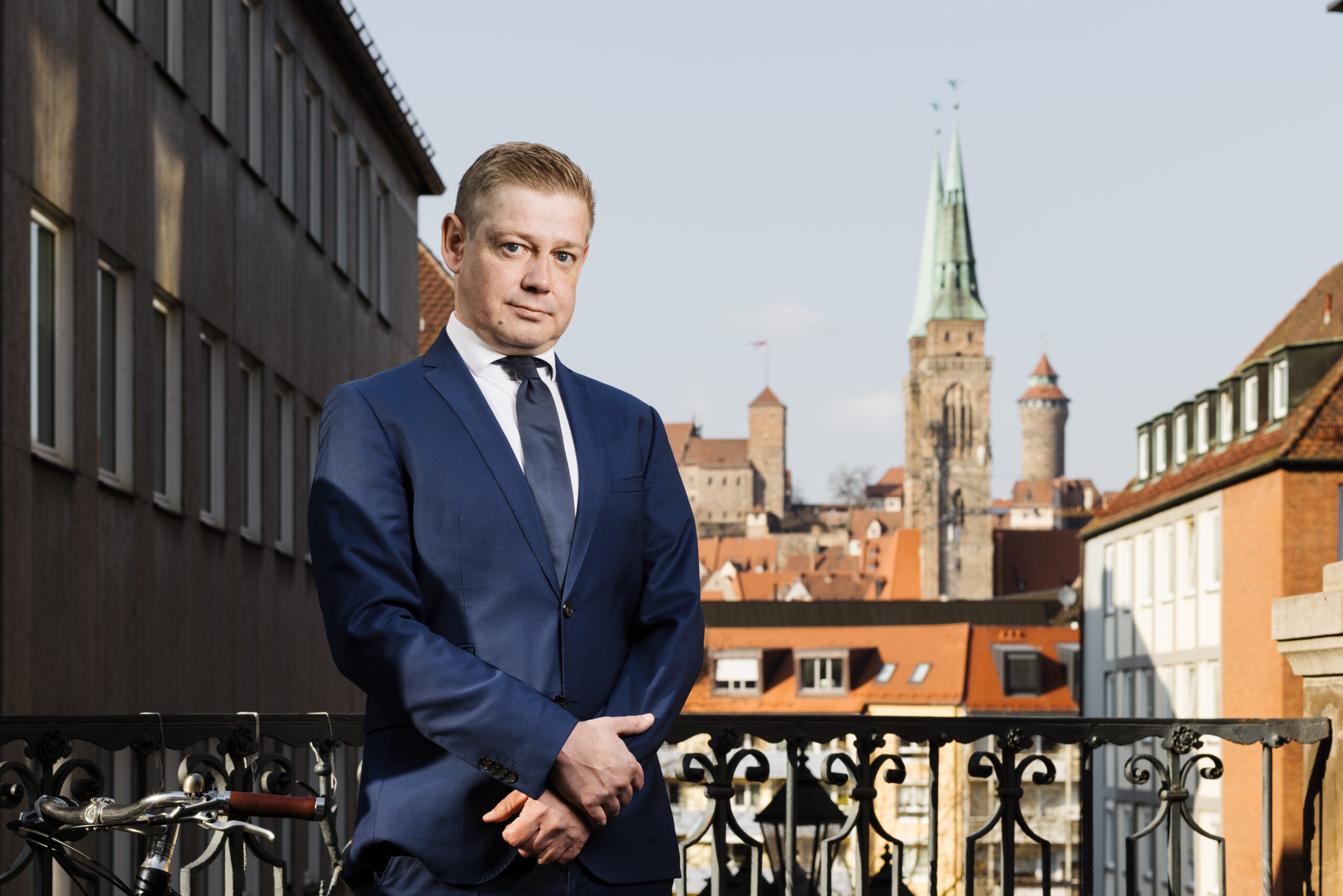 Fotografie Rechtsanwalt Kevin Nowack in schwarzem Mantel, Hemd und Krawatte, im Hintegrund Gebäude von Nürnberg